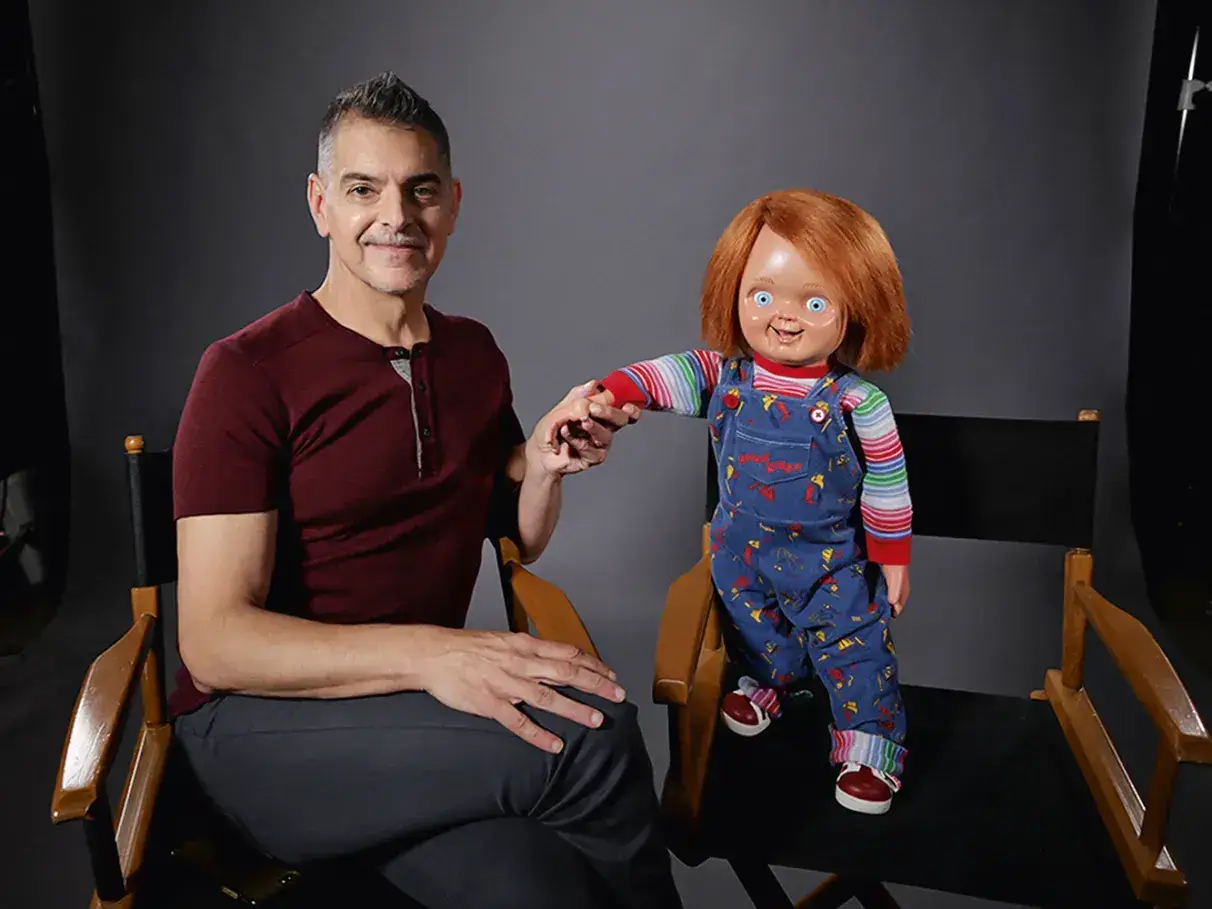 O Produtor Don Mancini está sentado em uma cadeira segurando a mão do boneco Chucky. Post O Retorno de Chucky ao Cinema.