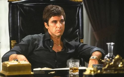 O personagem Scarface de Al Pacino está sentado em uma cadeira chique. Post Filmes Para Ver Pelo Menos Uma Vez no Ano.